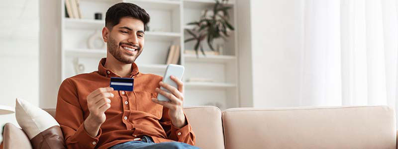 Hombre sonriente sentando en el sillón de su casa sosteniendo una tarjeta y el celular